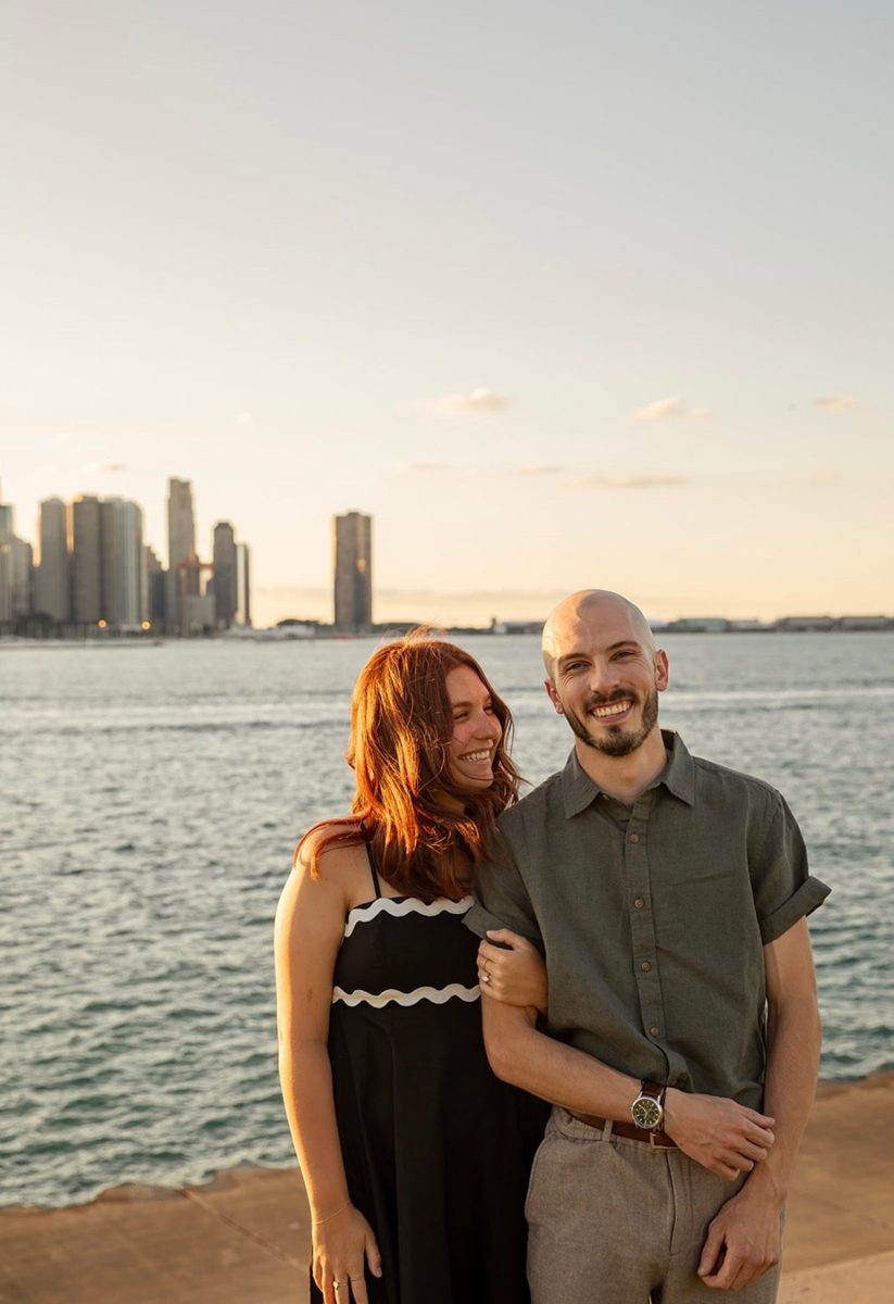 Kyle and Karli smiling arm in arm by the lake at sunset, with a city skyline behind them
