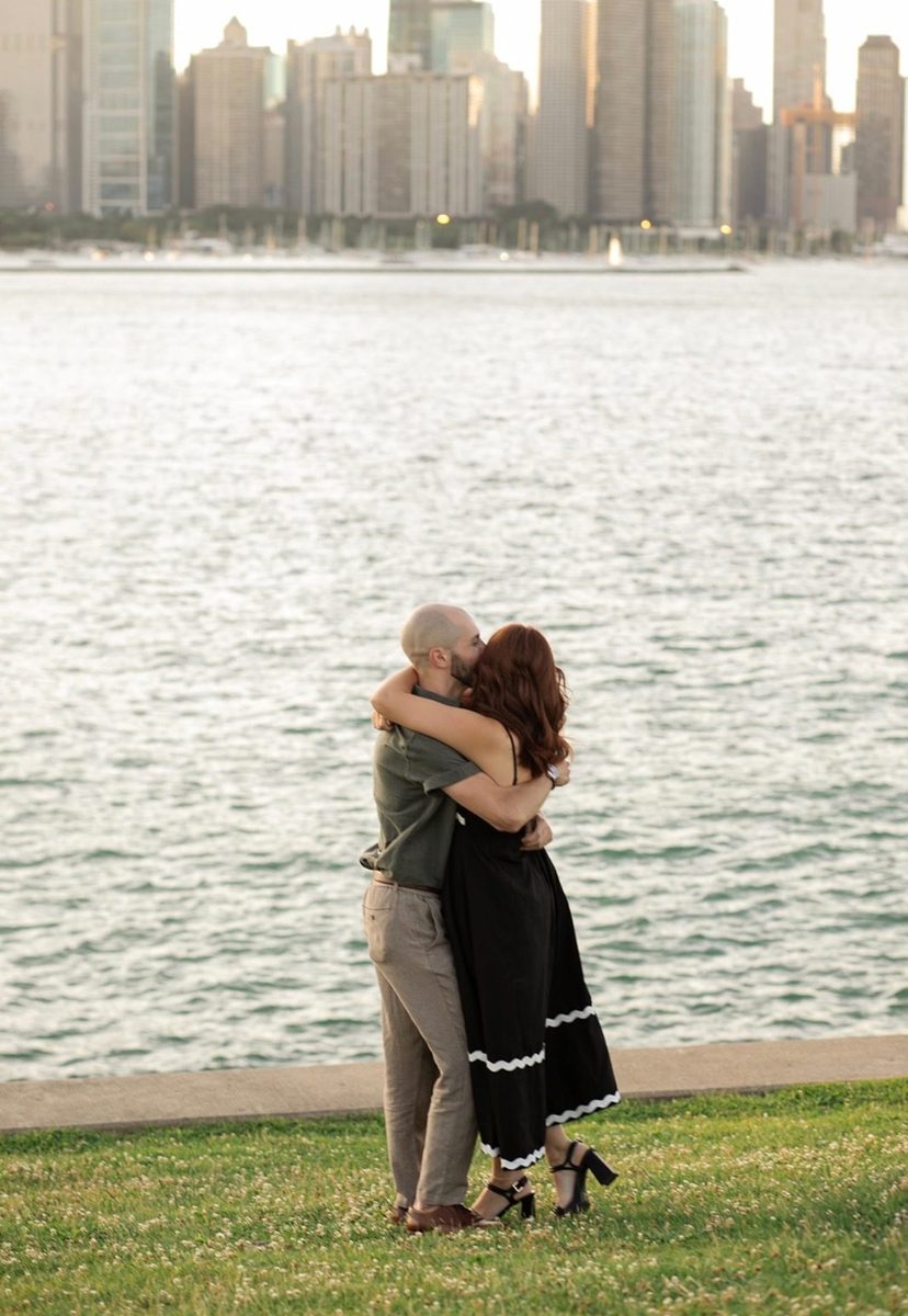Kyle and Karli embracing on the grass beside the lake, with the skyline across the water