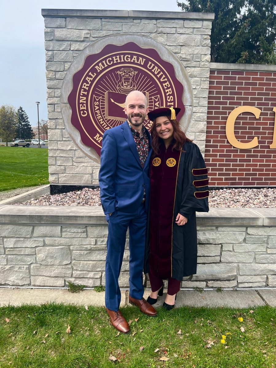 Kyle and Karli standing in front of a university sign, with Karli in her graduation regalia
