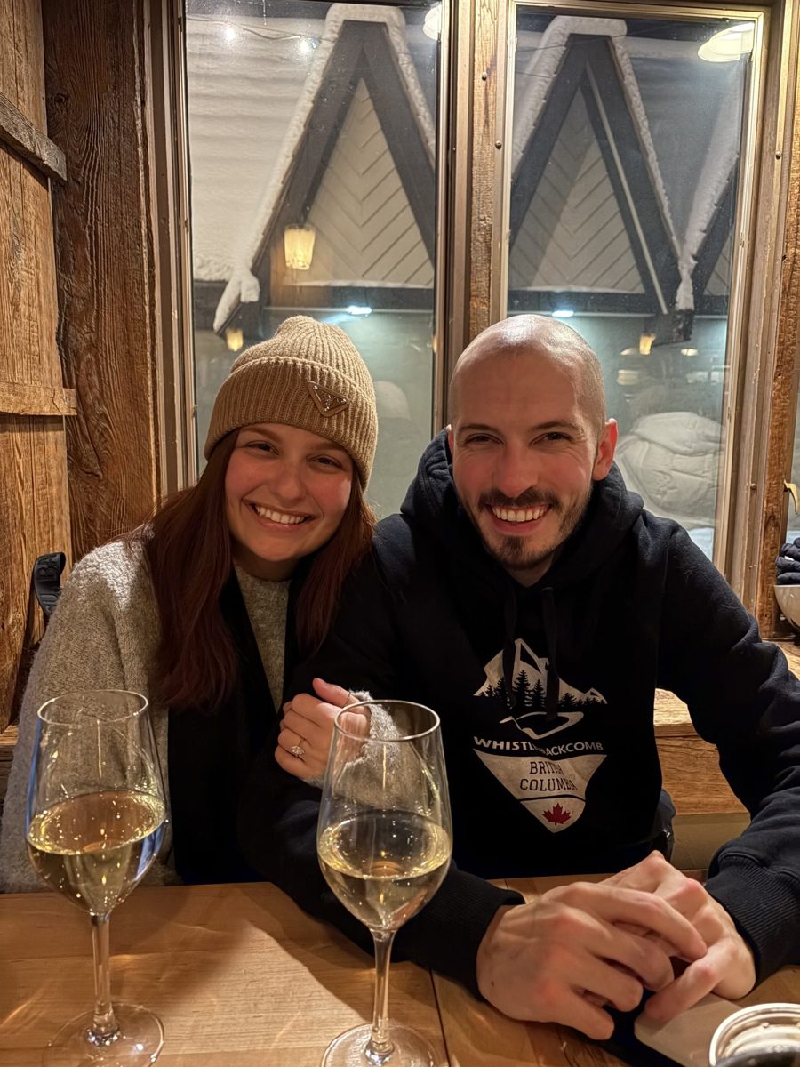 Kyle and Karli smiling at a wooden table with glasses of white wine