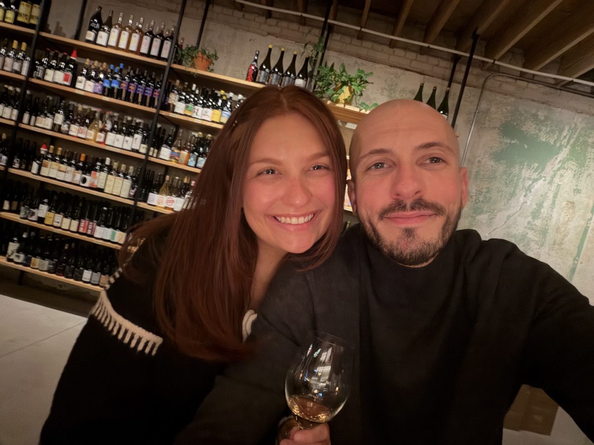 Kyle and Karli smiling close together with a glass of wine in front of a wall of wine bottles