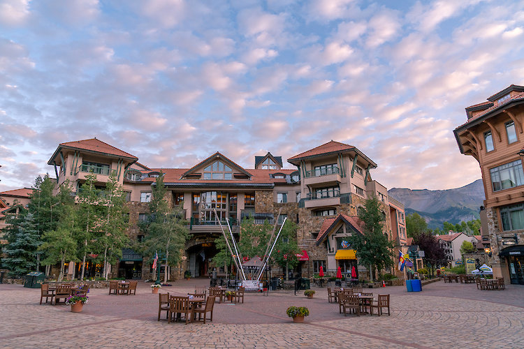 Mountain Village plaza with terra-cotta-roofed hotel buildings and aspens under a pink evening sky