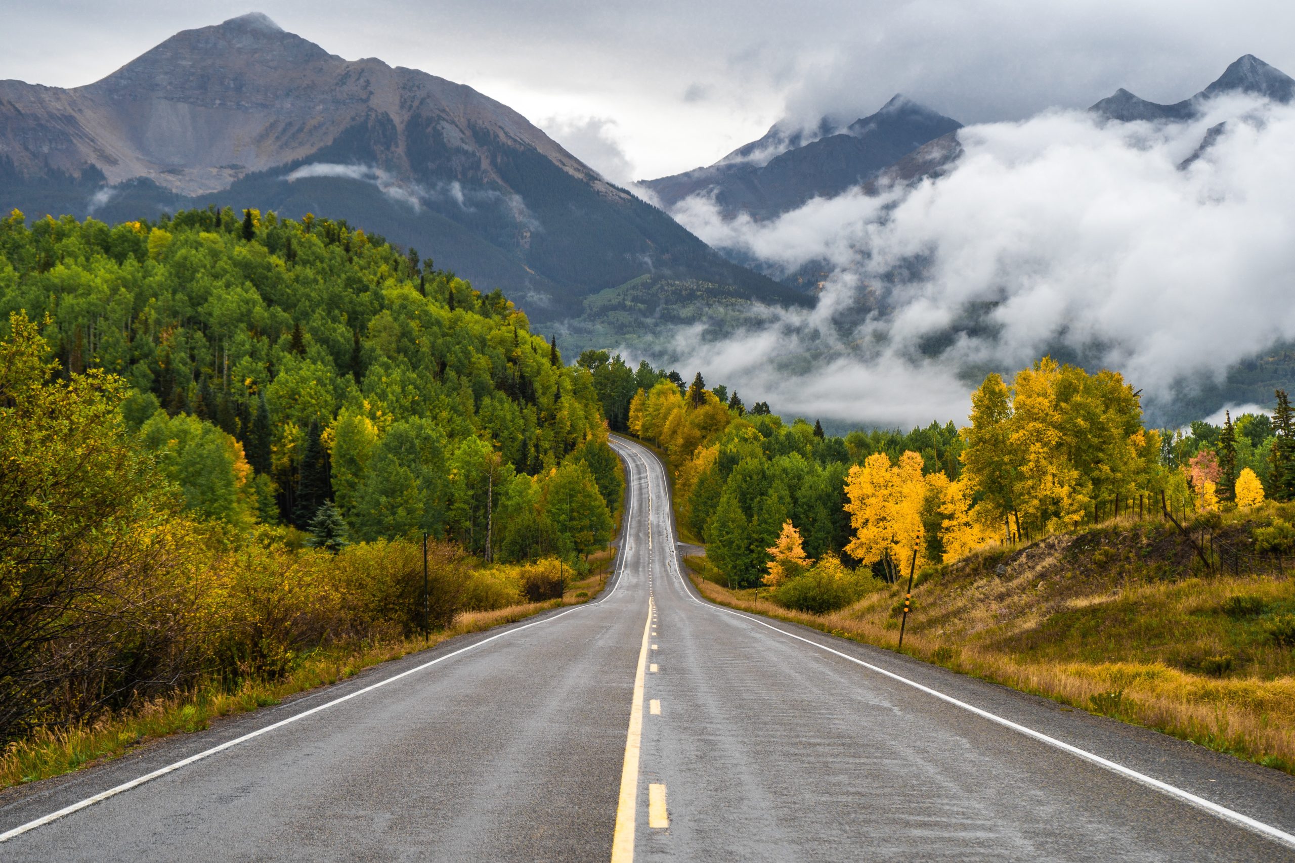 A two-lane mountain road through green and gold aspens toward cloud-wrapped San Juan peaks